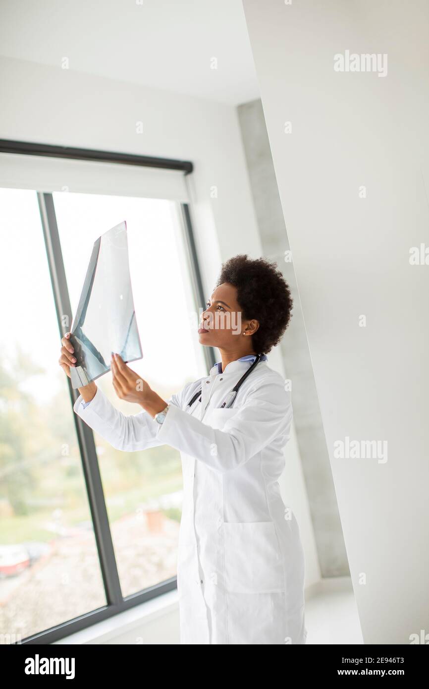 Donna Africana americana medico indossando camice bianco con stetoscopio in piedi dalla finestra in ufficio e guardando un'immagine radiografica Foto Stock