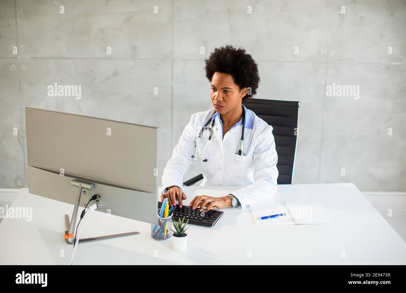 Donna Africana americana medico indossando camice bianco con stetoscopio seduta dietro la scrivania in ufficio Foto Stock