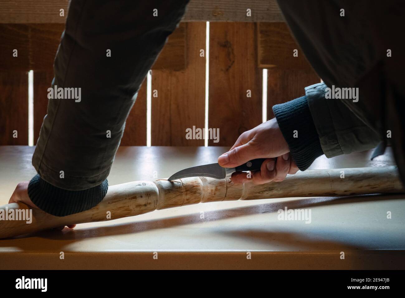 Processo di uomo che fa bastone di legno che cammina all'interno durante la quarantena. Bastone di legno intagliato sulla tavola usando il coltello Foto Stock