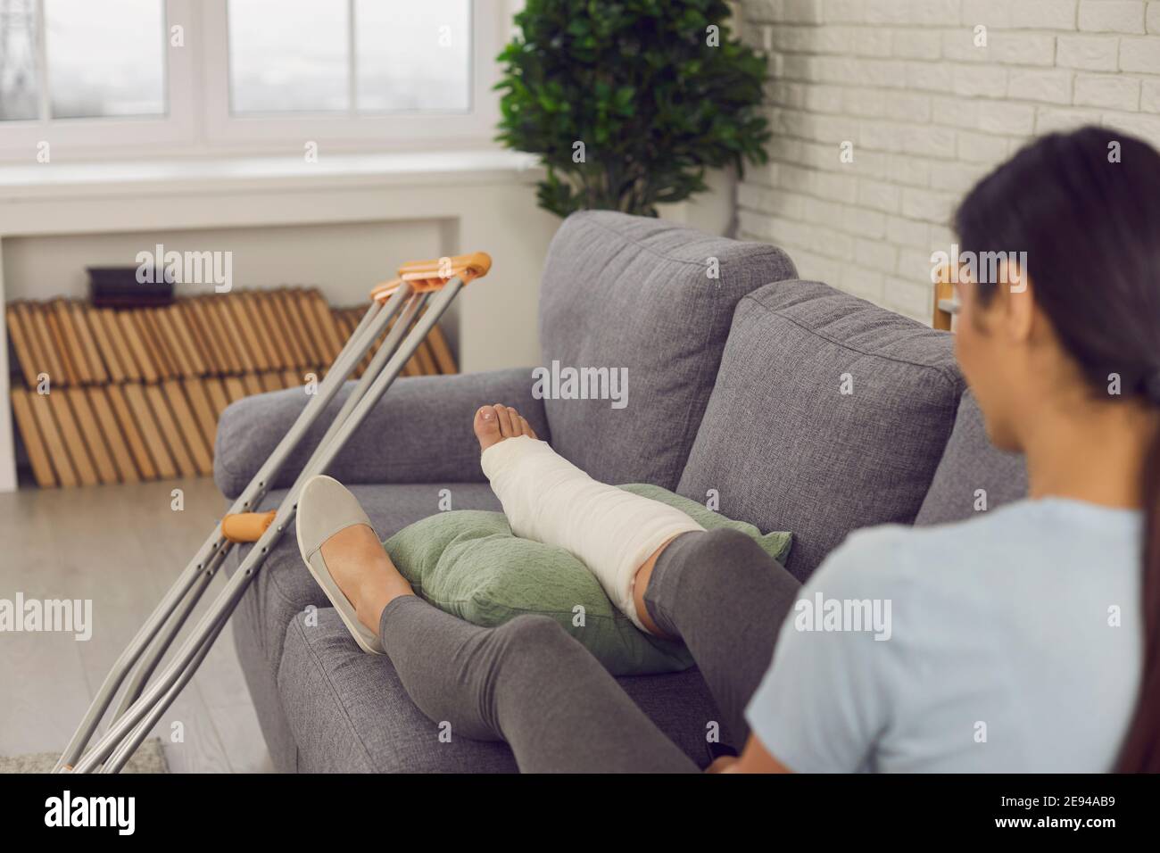 Paziente con gamba rotta in calco di gesso seduto sul divano a casa con le stampelle accanto Foto Stock
