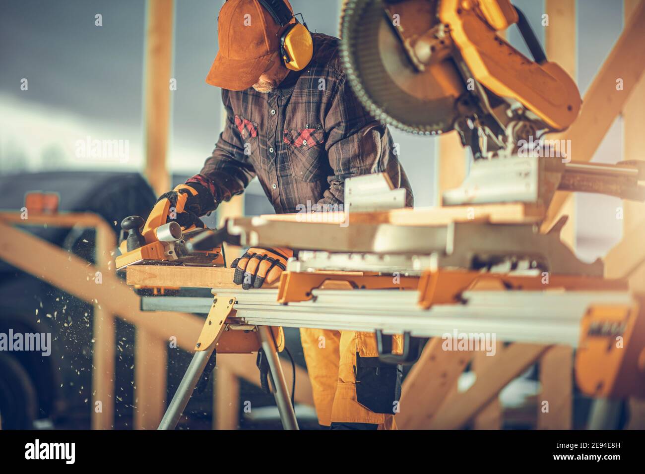 Caucasian Construction Worker nei suoi anni '40 con Noise Reduction Headphones Sulla sua testa che pianifica legno con la pialla elettrica all'interno di nuovo Costruito Skele di legno Foto Stock