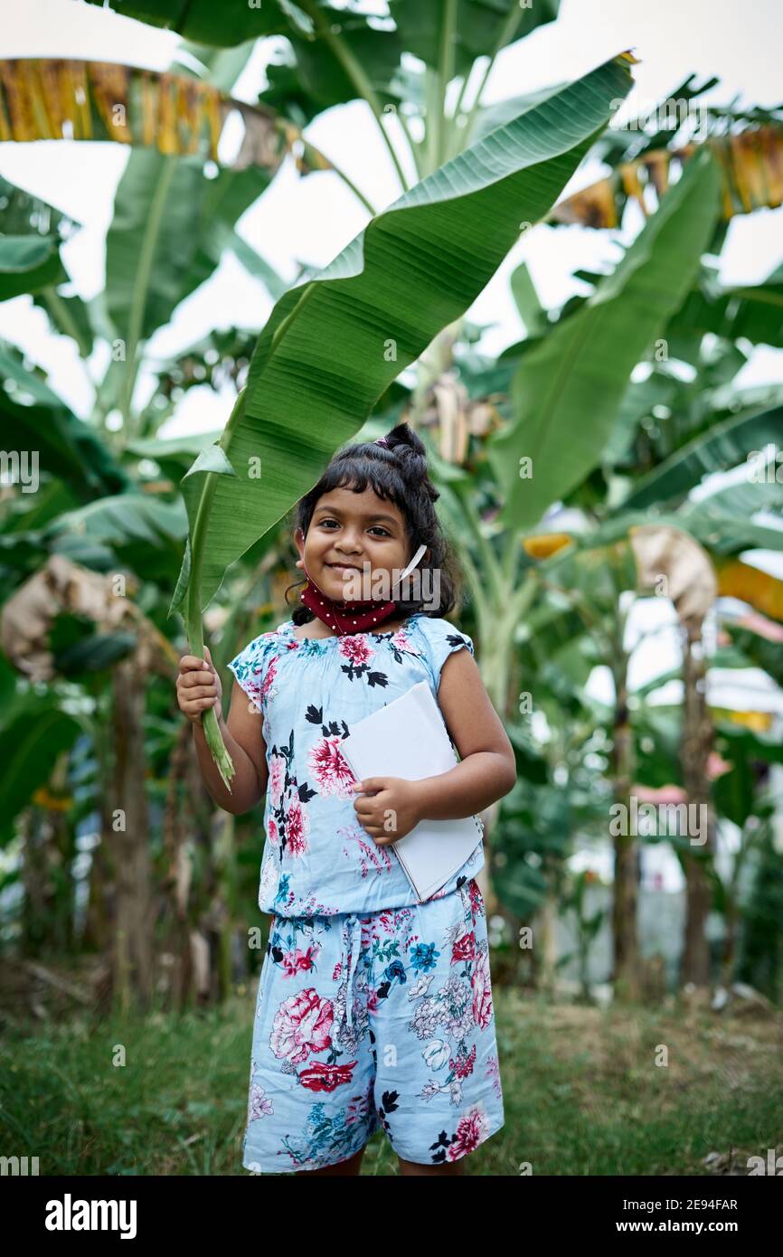 Piccola studentessa indossando maschera facciale e coprendo la testa con banana foglia andare a scuola Foto Stock