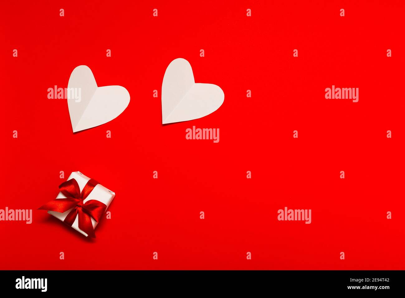 Composizione per San Valentino. Piccola scatola regalo bianca con nastro rosso e due cuori di carta bianca su sfondo rosso in stile piatto. San Valentino Foto Stock