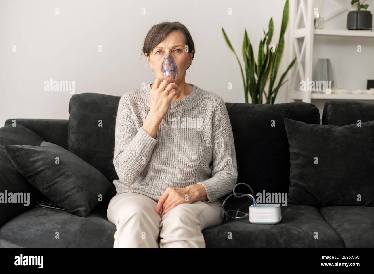 Donna anziana che usa l'inalatore per il trattamento dell'influenza e del freddo seduto sul divano a casa. Concetto di sanità e medicina Foto Stock