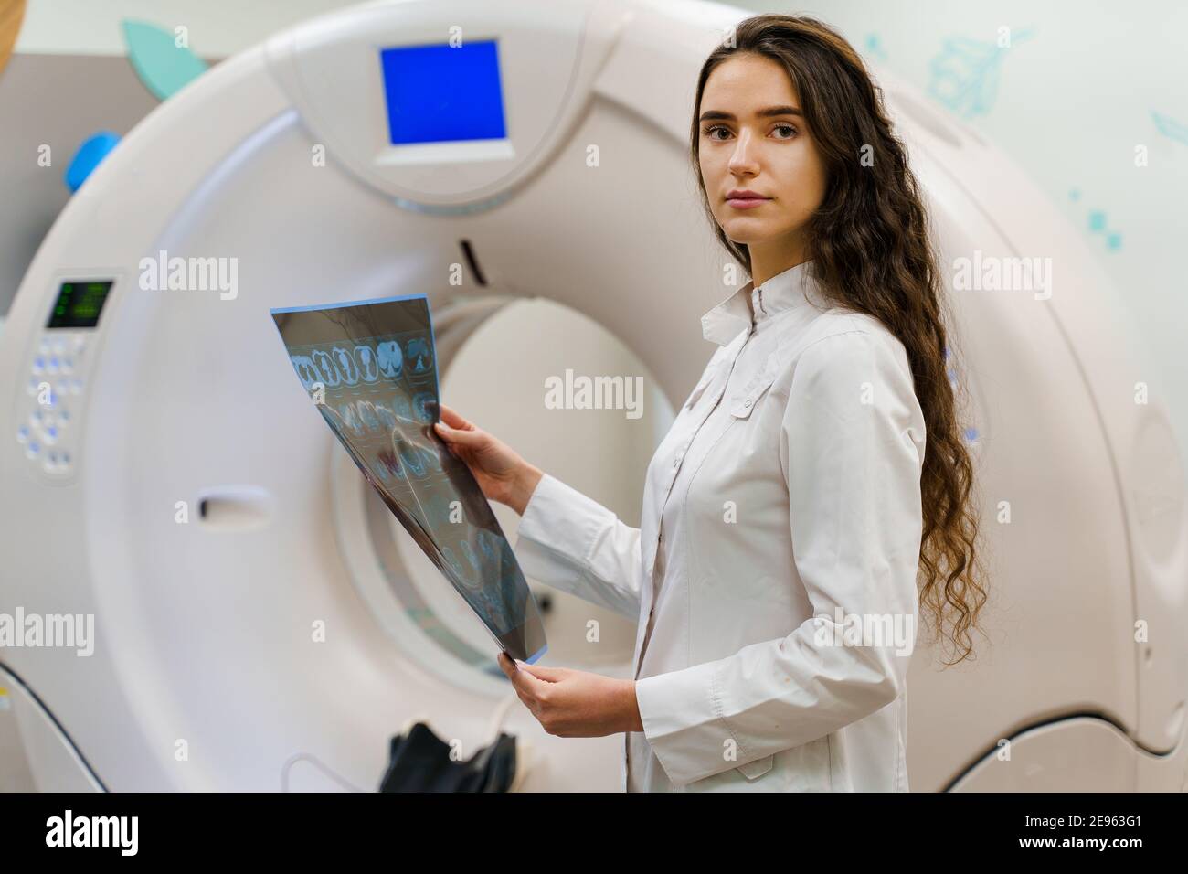 L'allievo medico guarda in macchina fotografica con l'immagine di x-ray del cervello del paziente nelle sue mani. La donna rimane davanti a tomografia di komputer. Giovane ragazza medica Foto Stock