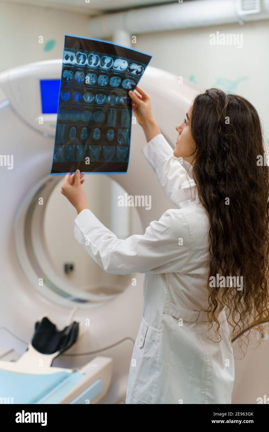 Immagine verticale dello studente medico che guarda l'immagine radiografica del cervello paziente vicino alla tomografia komputer. Giovane ragazza in clinica medica gotes nuovo e Foto Stock