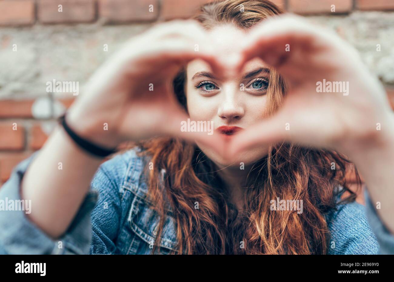 Ritratto di rosso arricciato capelli lunghi caucasici teen ragazza con labbra di rossetto rosso applicate con occhi blu sul mattone sfondo della parete che forma un cuore w Foto Stock