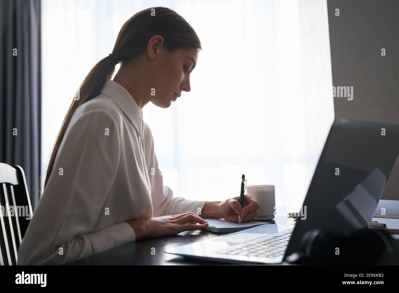 Vista laterale di un'affascinante signora che lavora a distanza su un computer portatile wireless mentre si siede al tavolo. Freelancer femminile in camicia bianca usando il computer portatile a ho Foto Stock