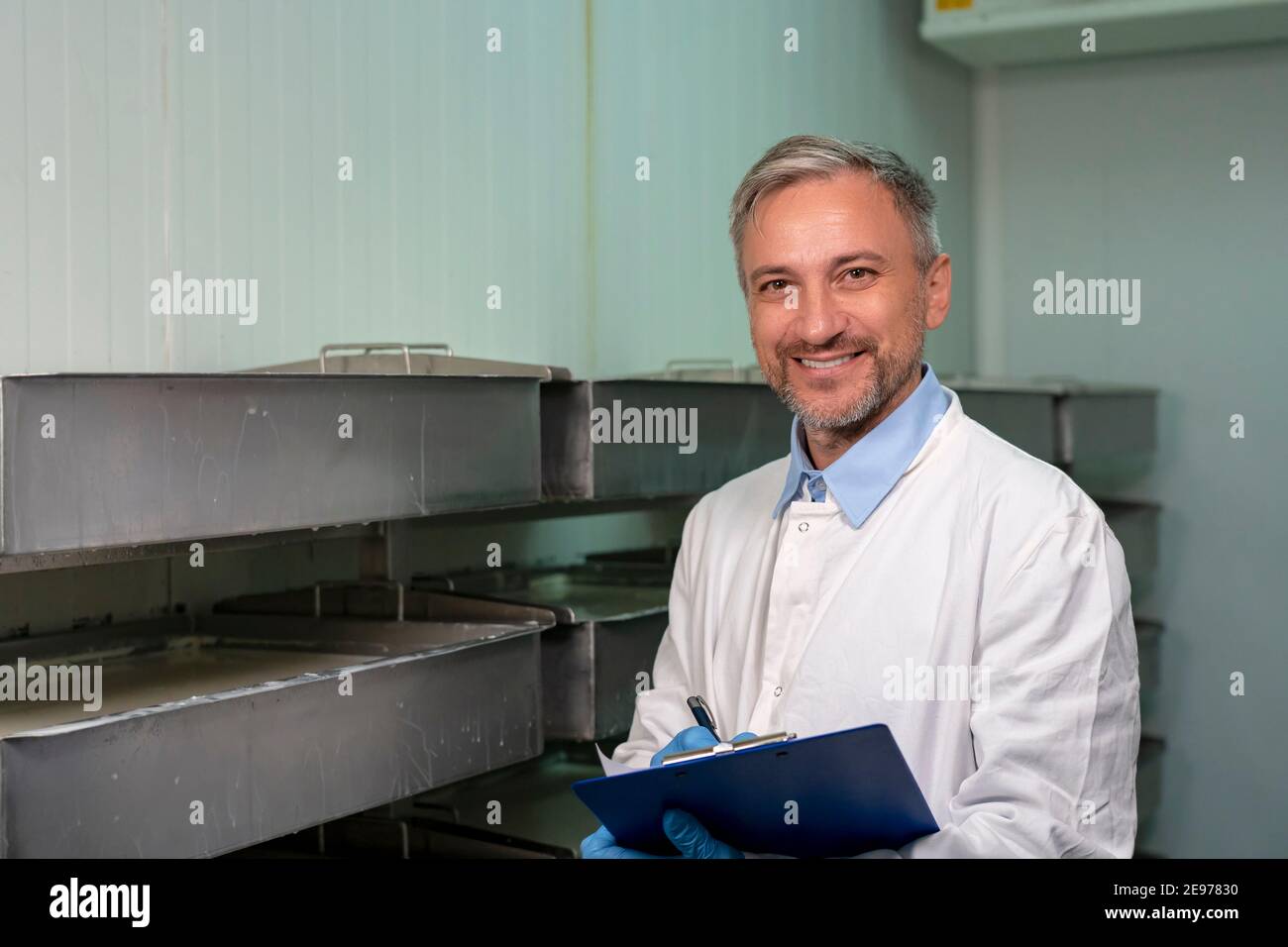 Happy Mature Man con Clipboard che indossa uniforme bianca e Guanti Blu, in piedi accanto alla linea di produzione del formaggio e guardando la fotocamera. Foto Stock
