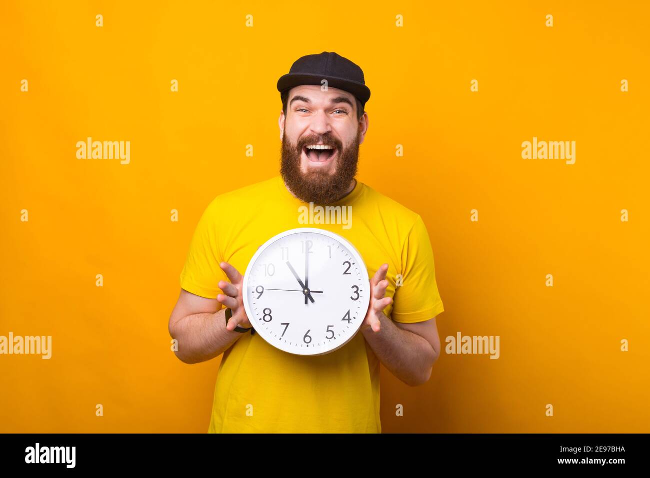 Immagine di un uomo eccitato che tiene un orologio vicino ad una parete gialla sorridente . Foto Stock