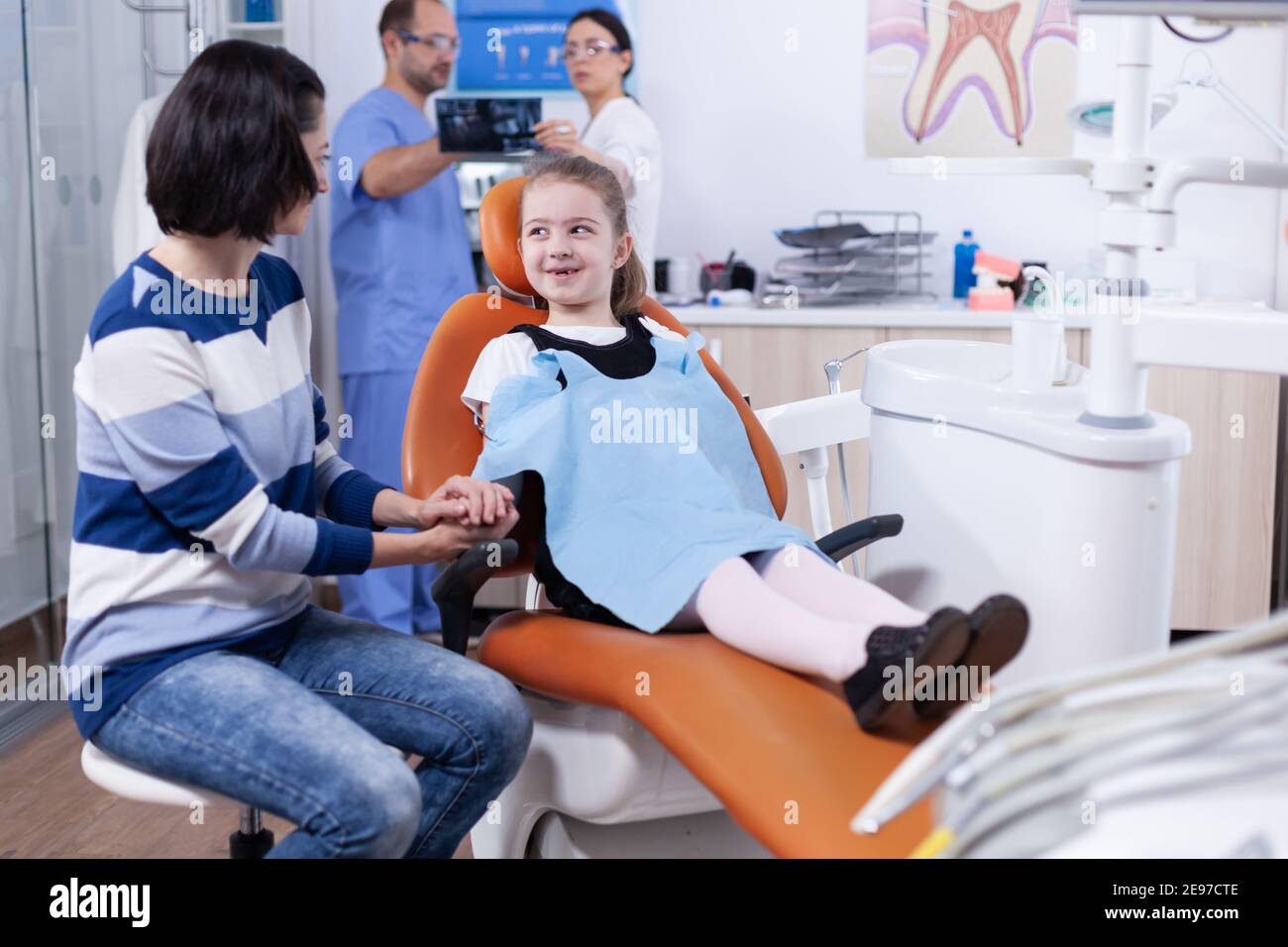 Bambina con dente mancante che indossa un bib dentale in attesa di esame in ufficio dentista. Bambino con la madre durante il controllo dei denti con stomatolog seduta sulla sedia. Foto Stock