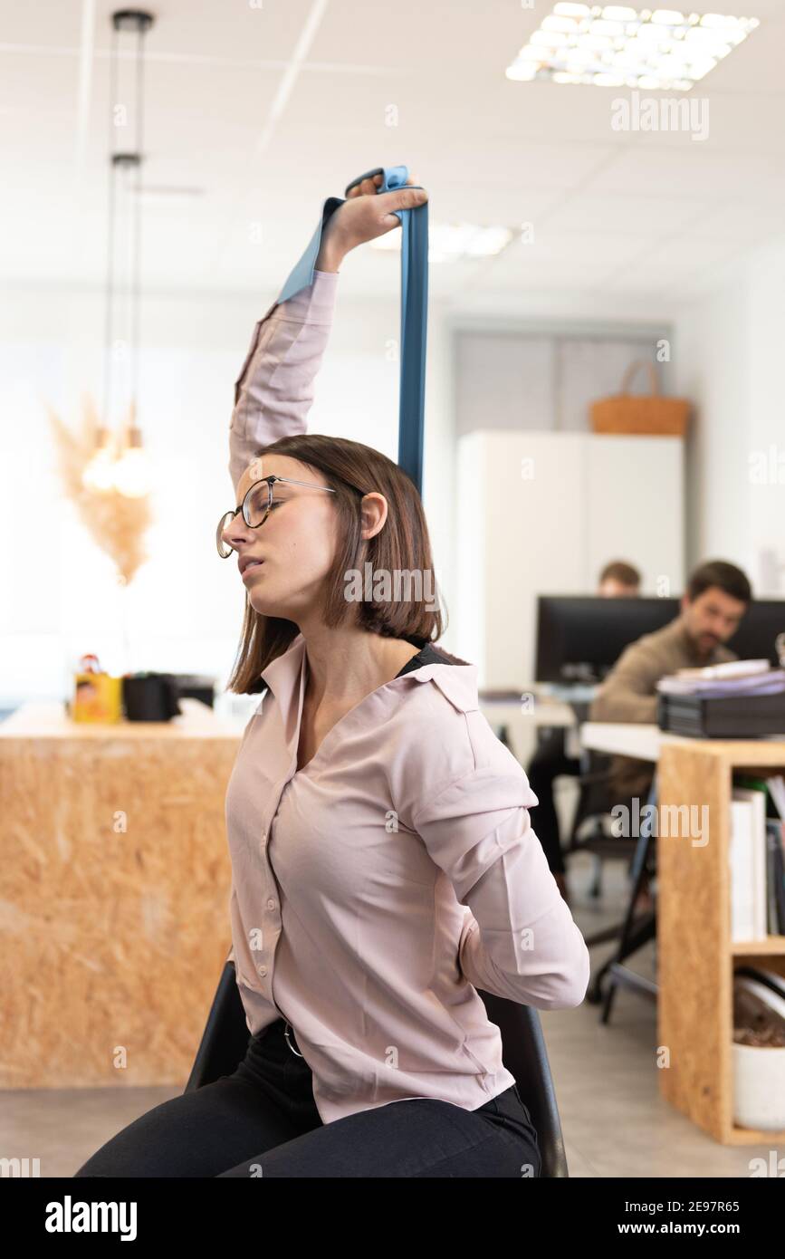 Stile di vita sano nel concetto di luogo di lavoro. Donna con mal di schiena che si allunga e rilassa la schiena con una fascia in gomma pilates in ufficio. Foto Stock