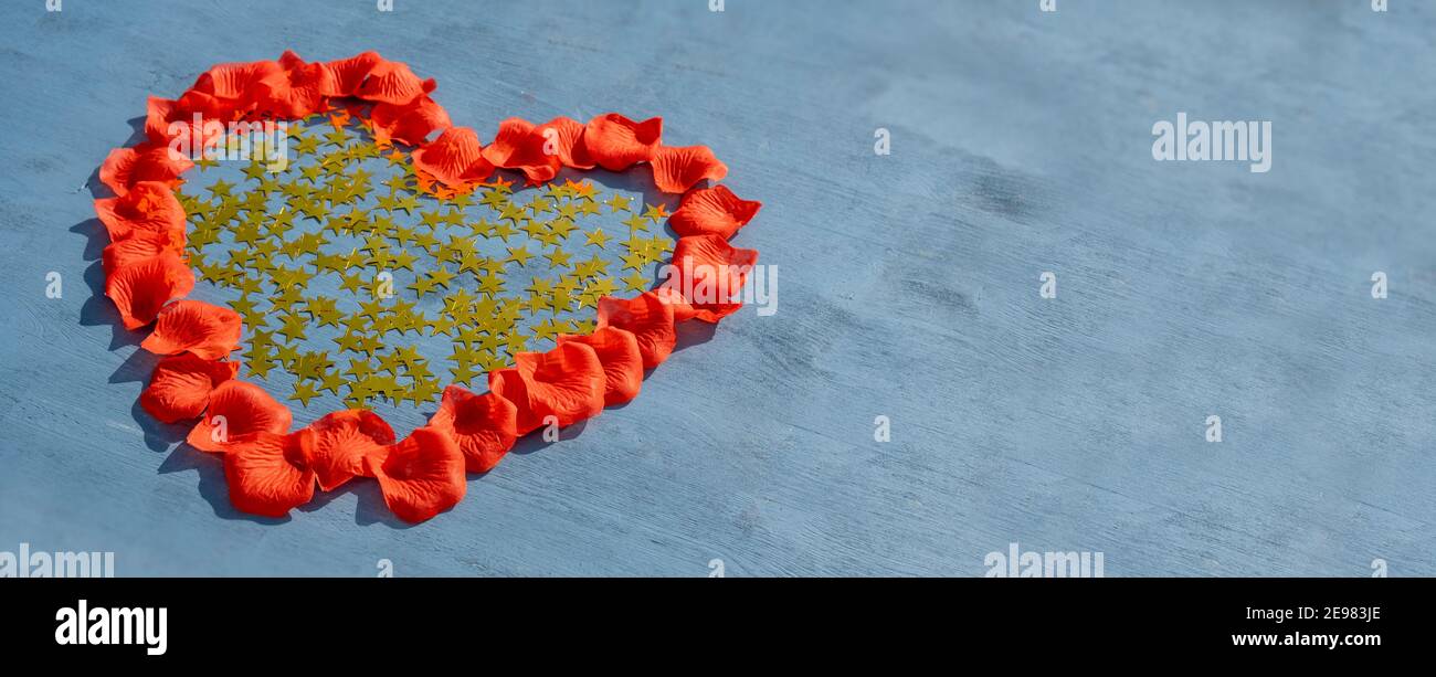 Concetto di giorno Love and Valentines: Vista dall'alto su una forma a cuore fatta di foglie di rosa rossa. Il cuore è pieno di stelle metalliche d'oro su legno blu Foto Stock