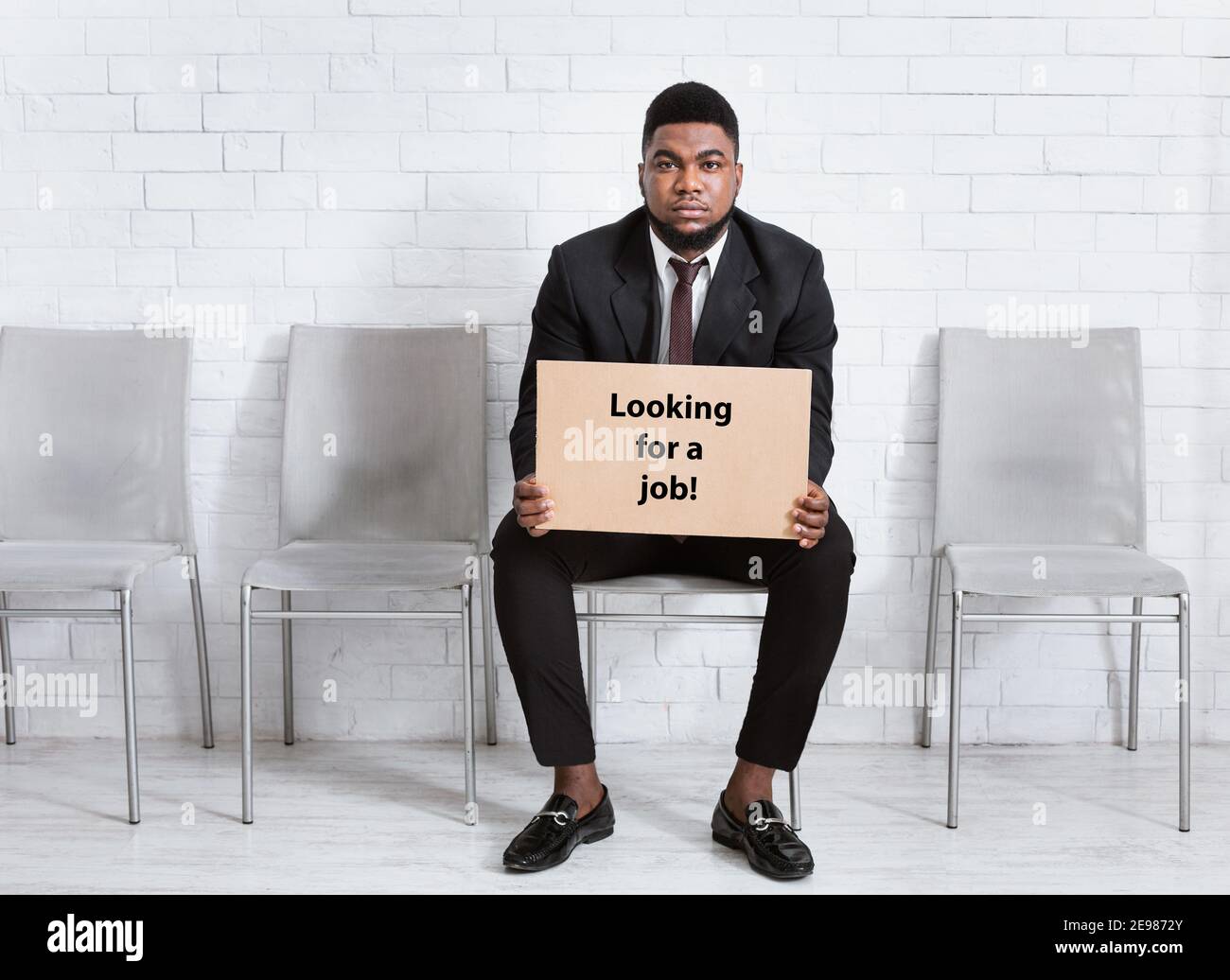 Uomo afro-americano in tuta formale tenuta IN CERCA DI UN CARTELLO DI LAVORO, in attesa di un colloquio di lavoro nella lobby dell'ufficio, spazio libero Foto Stock