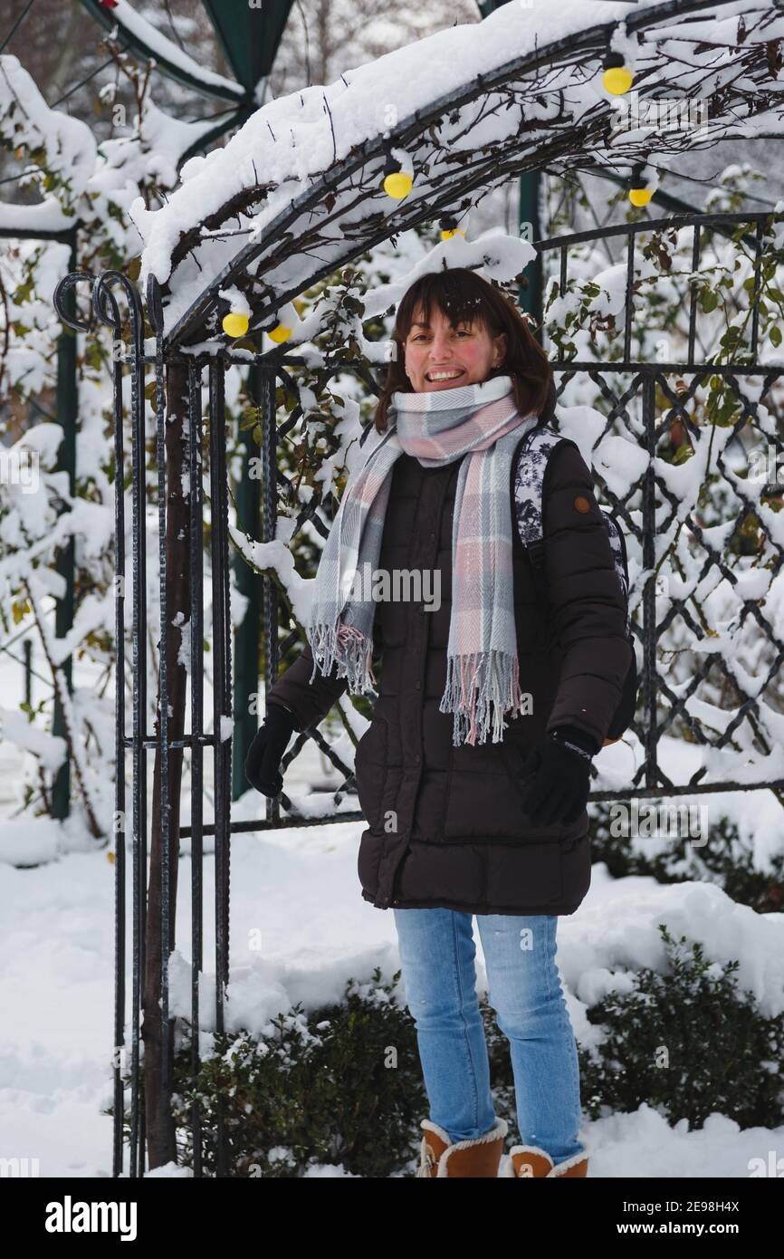 Giovane donna sorridente con giacca lunga marrone e sciarpa rosa grigia in un parco invernale innevato. Inverno innevato, giorno gelido Foto Stock