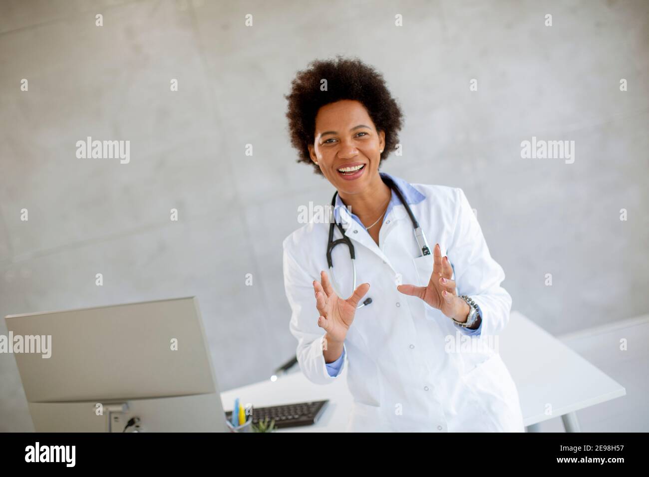 Donna Africana americana medico indossando camice bianco con stetoscopio in piedi dalla scrivania in ufficio Foto Stock
