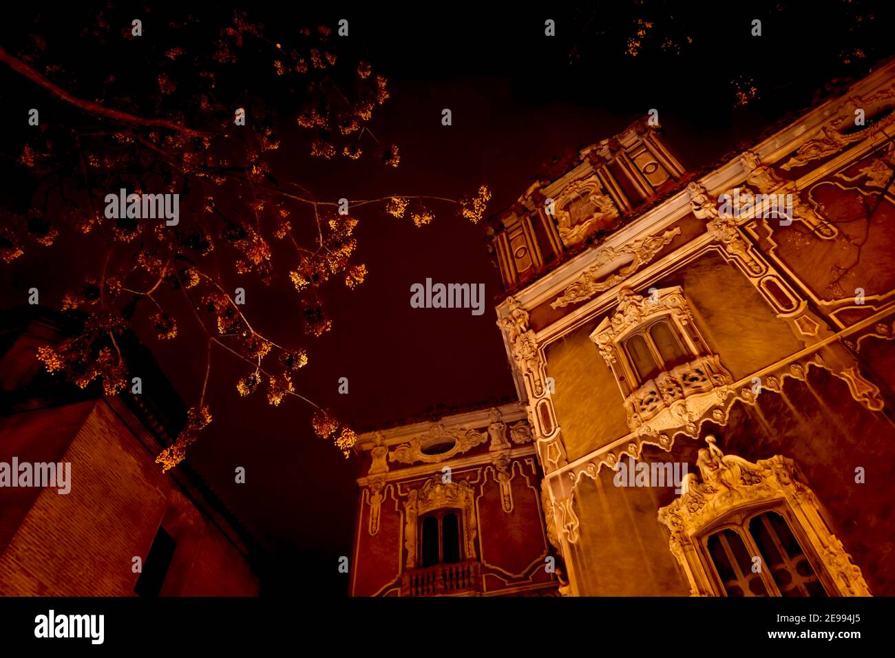 Facciata barocca di 'Gonzalez Marti' il Museo Nazionale della Ceramica E arti decorative durante la notte illuminata di arancio con fiori sfocati albero in sp Foto Stock