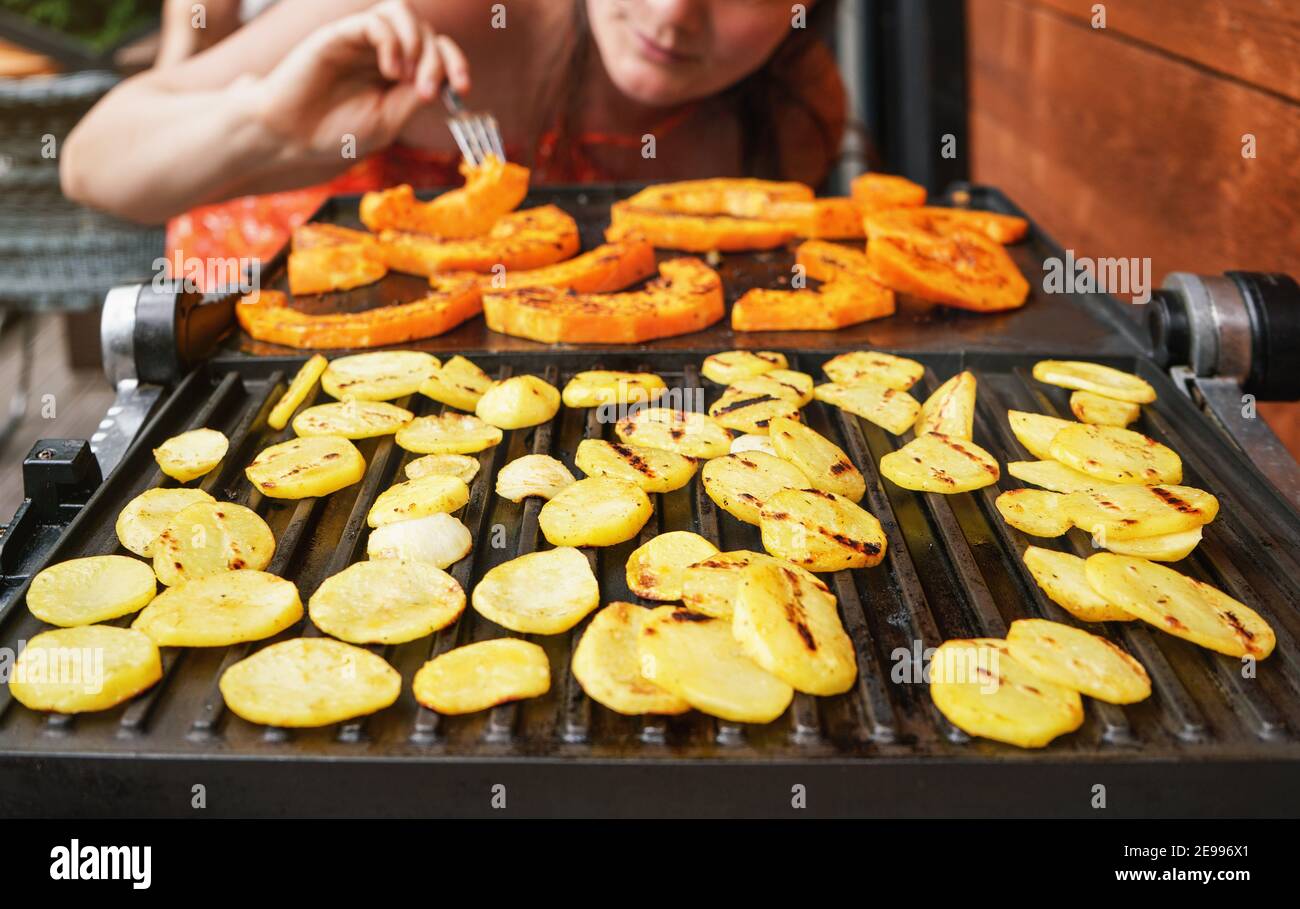 Patatine e pezzi di zucca butternut grigliati sulla griglia elettrica, concentrarsi su cerchi di patate brillanti, giovane donna sfocata in background Foto Stock