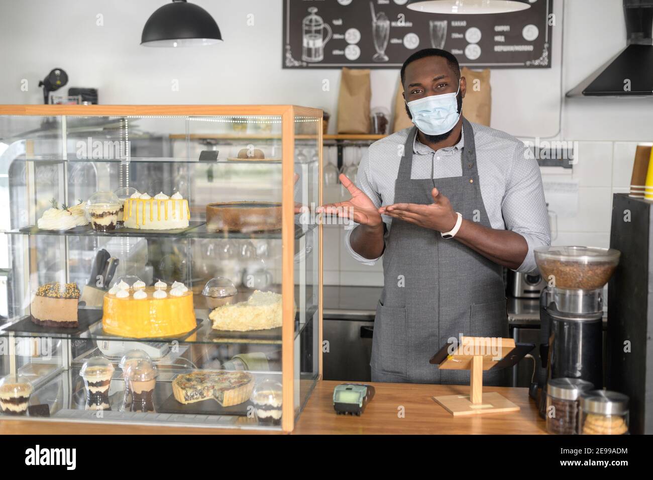 Un cameriere afroamericano maschio al bancone che indossa una maschera medica punta a pasticceria fresca e torte sulla vetrina. Una misura protettiva per il caffè Foto Stock