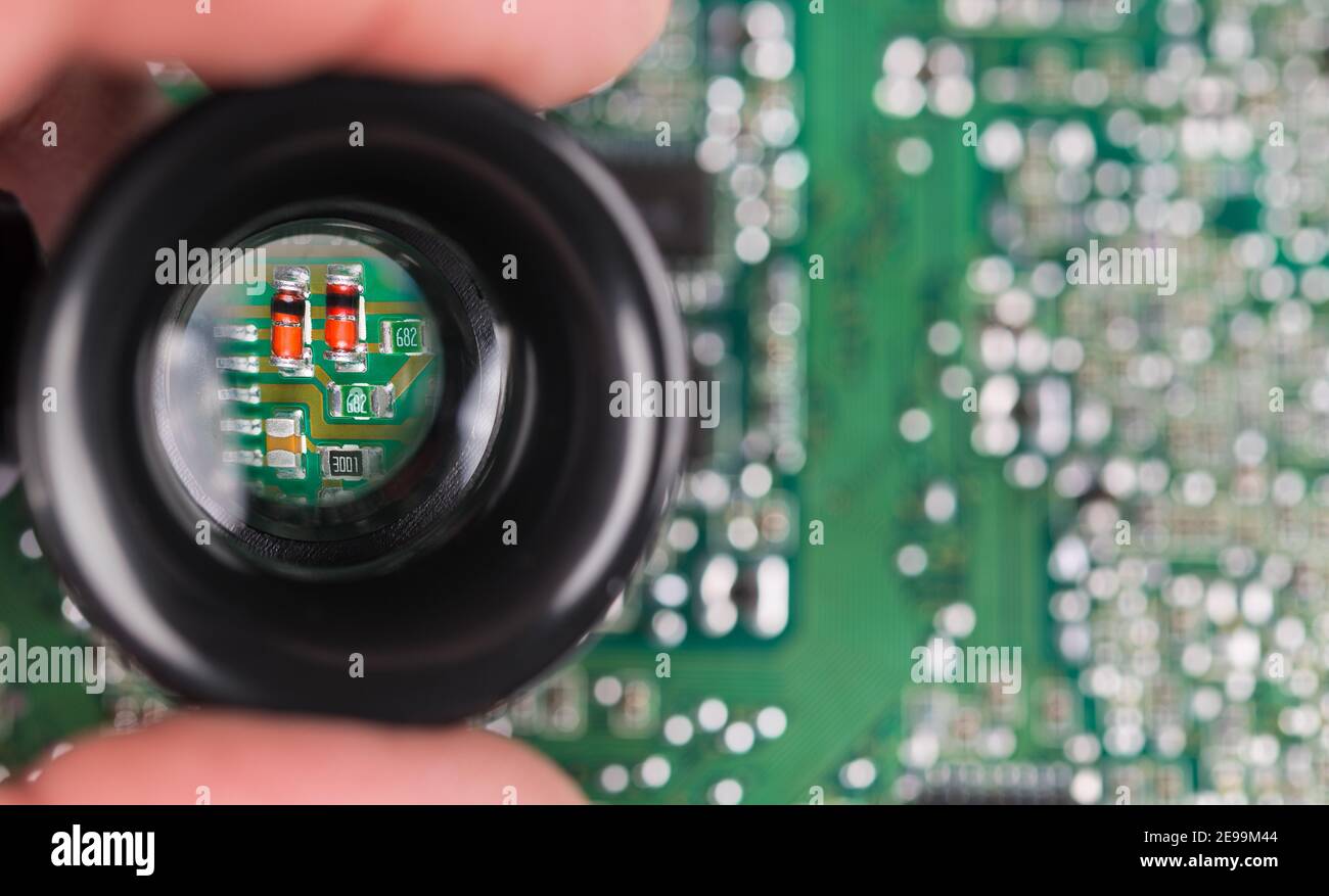 Piccoli componenti elettronici con feritoie nere su sfondo PCB verde. Primo piano di diodi a semiconduttore rossi ingranditi, resistori o condensatori nella lente manuale. Foto Stock