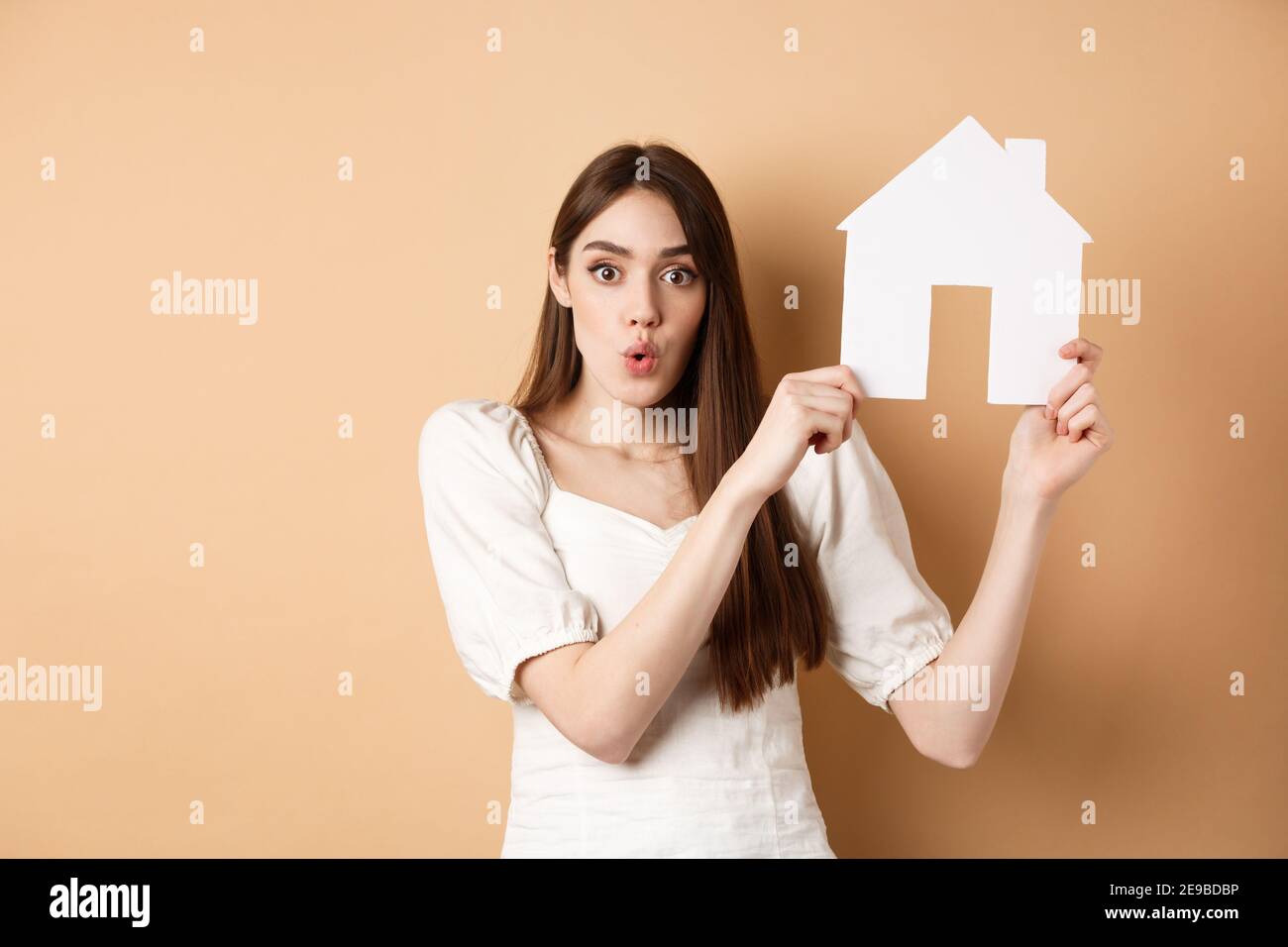 Immobiliare. Donna eccitata che mostra il taglio della casa di carta e che dice WOW con faccia stupita, proprietà d'acquisto, in piedi su sfondo beige Foto Stock