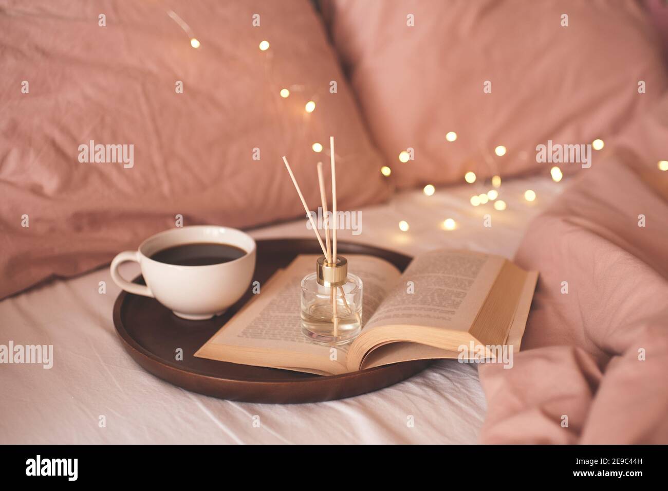 Tazza di caffè con libro di carta aperto su vassoio di legno con fragranza domestica profumata liquida nel letto sopra le luci incandescenti si avvicinano allo sfondo. Atmosfera accogliente Foto Stock