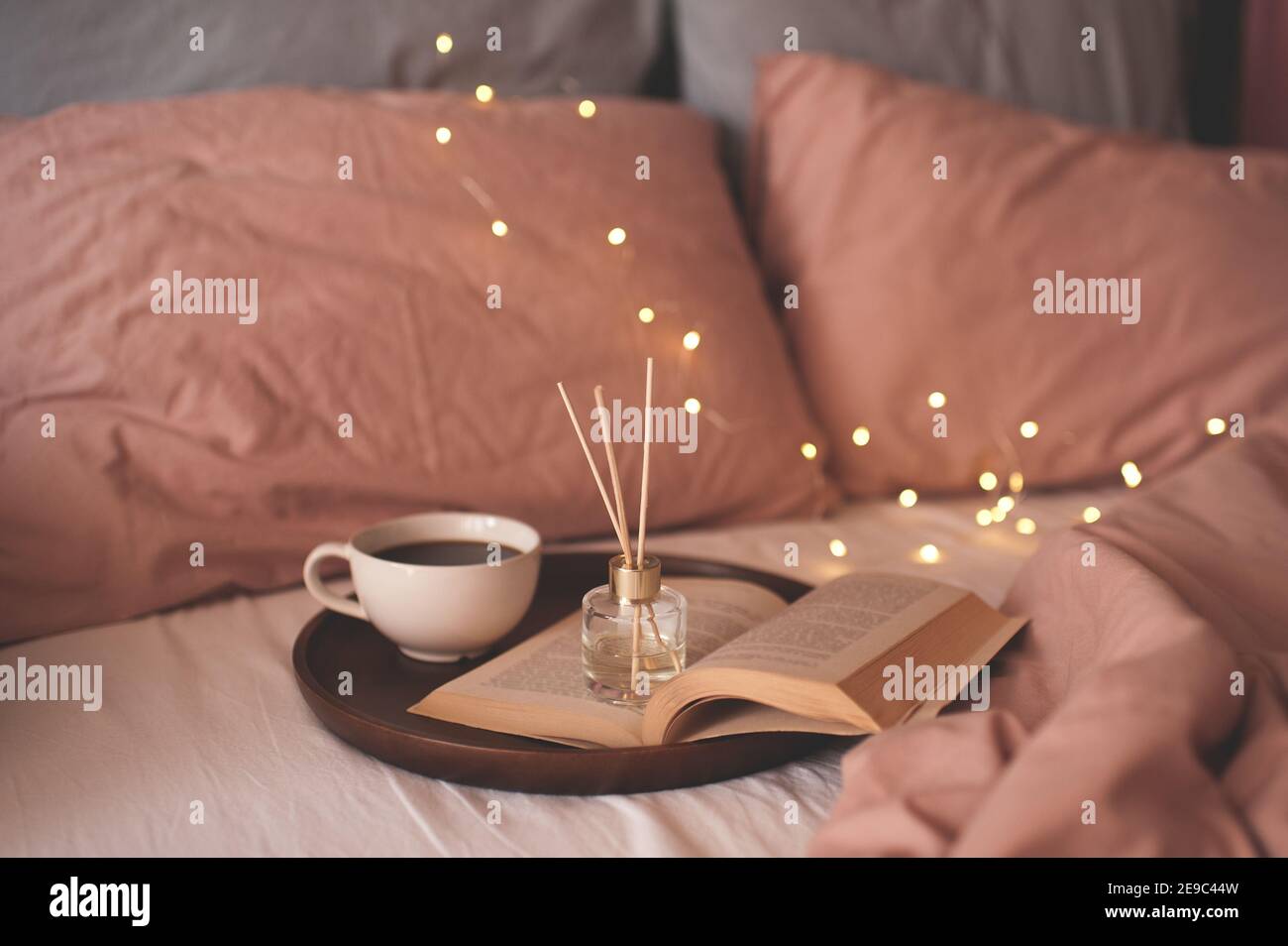 Tazza di caffè con libro di carta aperto su vassoio di legno con fragranza domestica profumata liquida nel letto sopra le luci incandescenti si avvicinano. Atmosfera accogliente. Goo Foto Stock