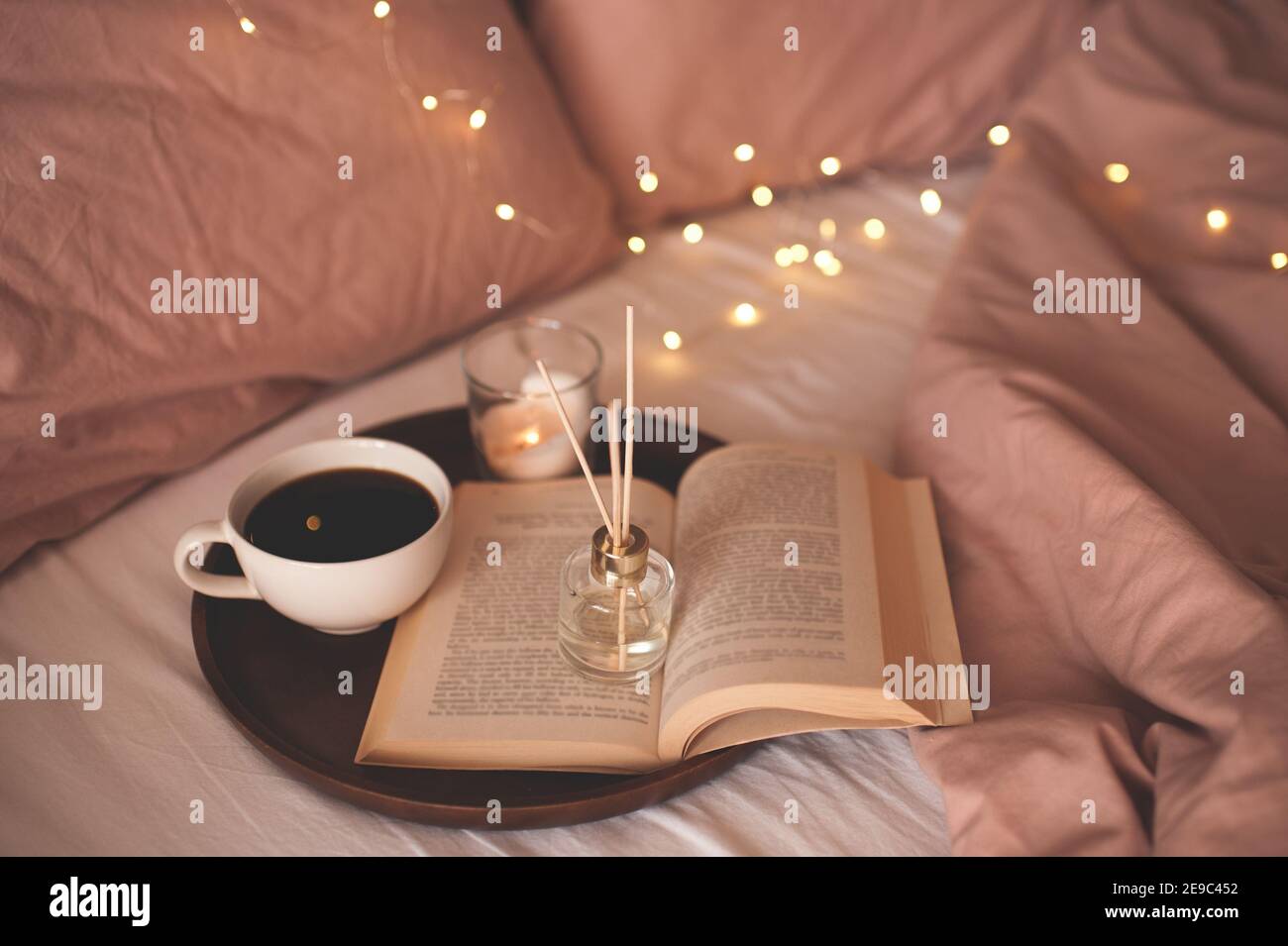Tazza di caffè con libro di carta aperto su vassoio di legno con fragranza domestica profumata liquida nel letto sopra le luci incandescenti si avvicinano allo sfondo. Atmosfera accogliente Foto Stock