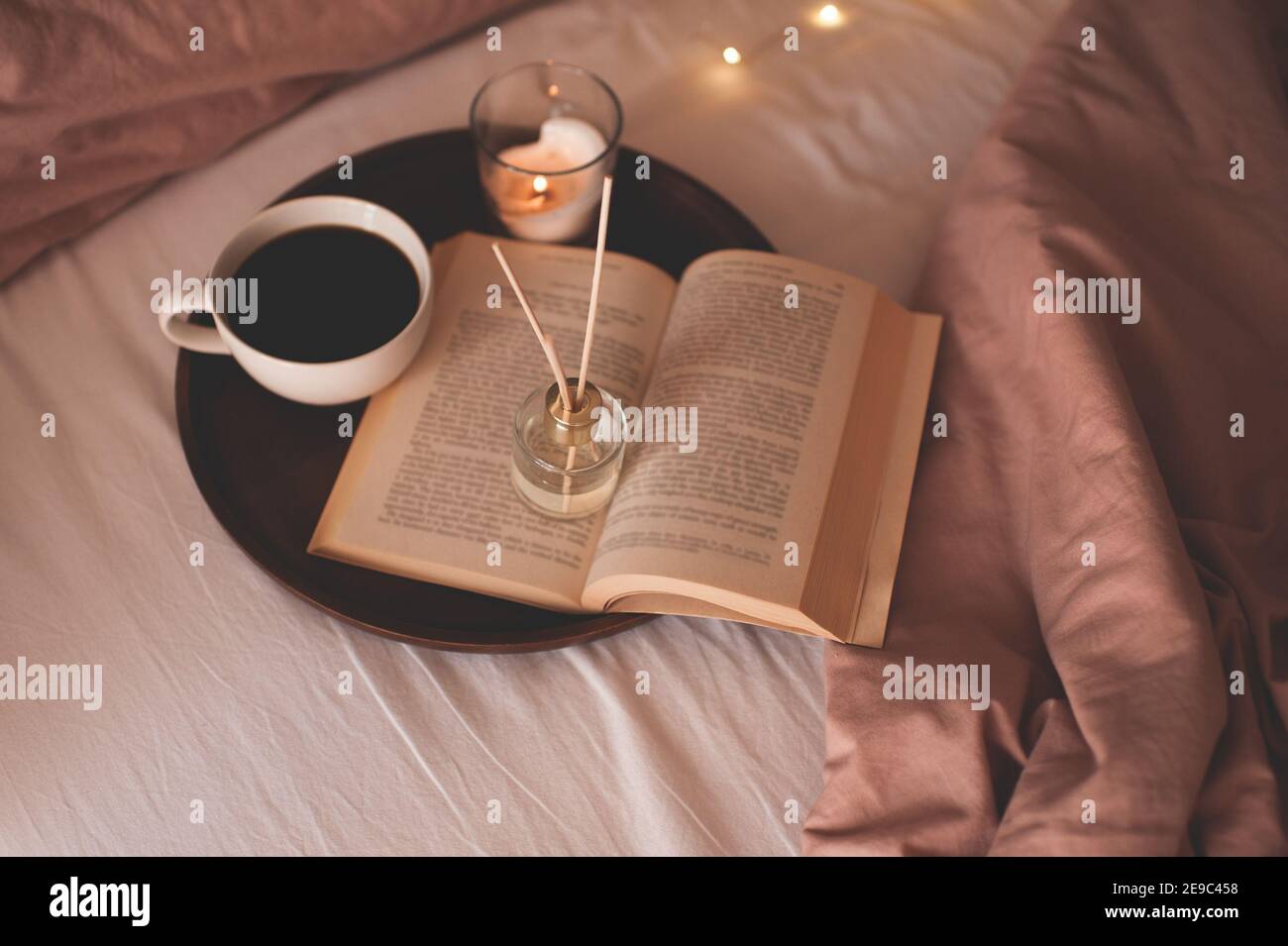 Tazza di caffè con libro di carta aperto su vassoio di legno con fragranza domestica profumata liquida nel letto sopra le luci incandescenti si avvicinano. Atmosfera accogliente. Goo Foto Stock