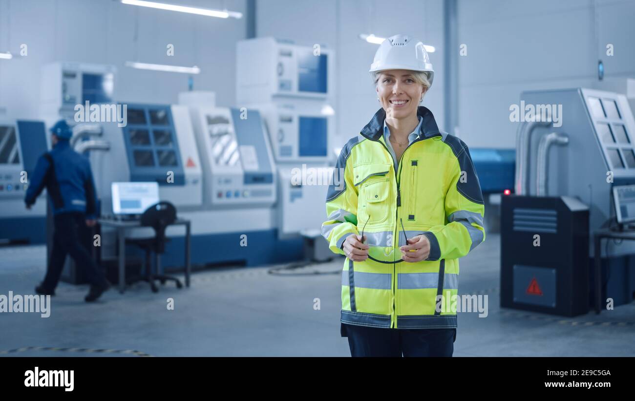 Bella femmina sorridente ingegnere indossare il giubbotto di sicurezza e Hardhat sostiene occhiali di sicurezza. Donna professionale che lavora nella moderna fabbrica di produzione Foto Stock