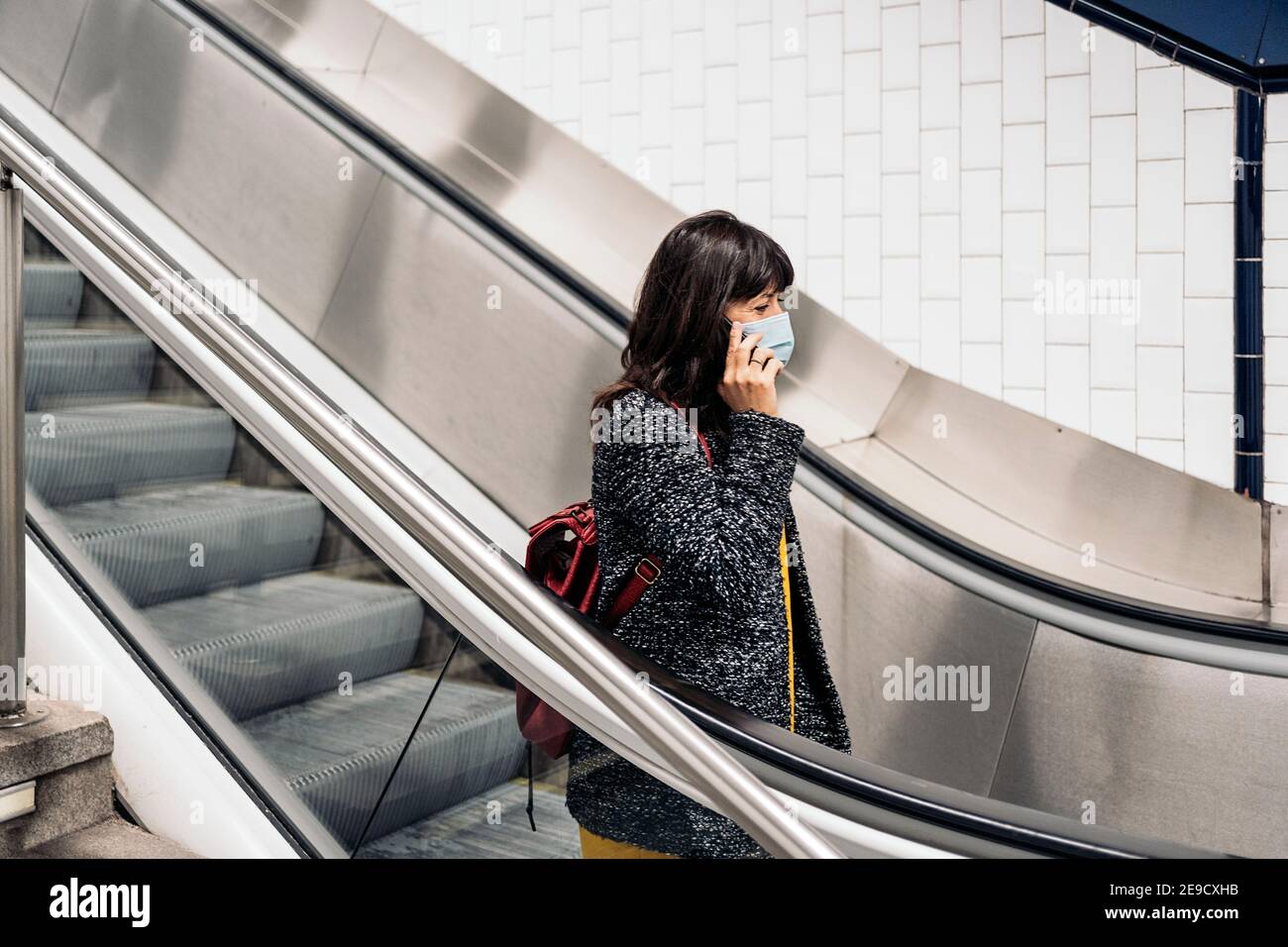 Foto di scorta di donna felice che indossa la maschera che parla al telefono mentre si sta in piedi nelle scale mobili. Foto Stock