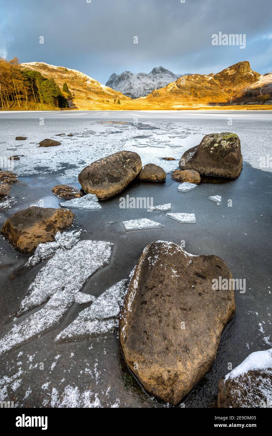 Un Blea Tarn ghiacciato con Langdale Pikes innevato sullo sfondo ripreso in una fredda mattina invernale con luce dorata e drammatiche nuvole nel cielo. Lago Foto Stock