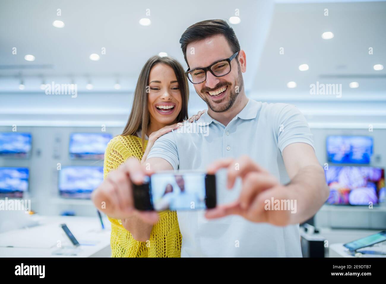 Coppia che fa selfie nel negozio di tecnologia. Negli schermi TV bacground. Foto Stock