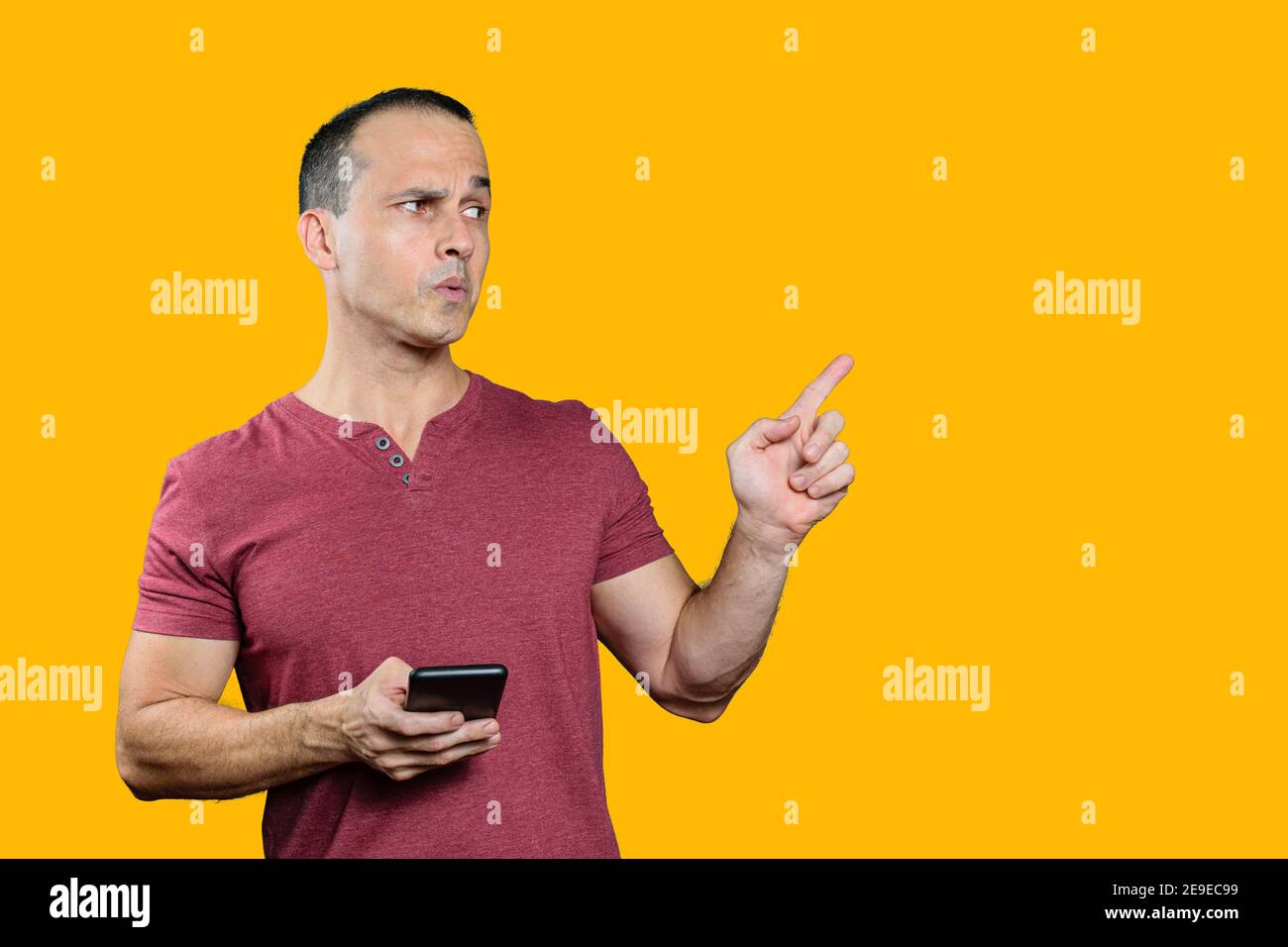 Uomo brasiliano maturo che tiene uno smartphone, puntando il dito a sinistra e facendo un volto. Sfondo arancione. Foto Stock