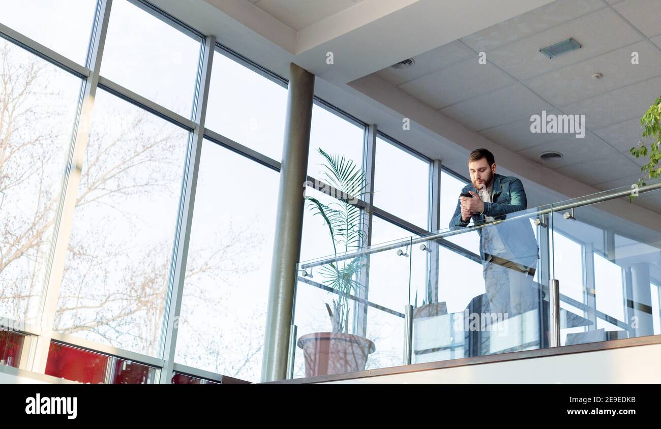 L'uomo della barba caucasica sta chiacchierando testando sullo smartphone in un moderno ufficio urbano di coworking sul balcone di vetro vicino alla finestra. Freelance hanno un telefono cellulare Foto Stock