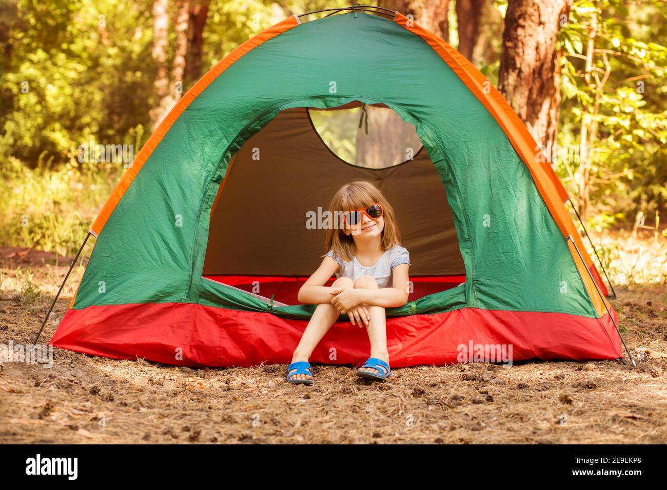 Bambina in occhiali da sole sorride e guardando la macchina fotografica che gioca in tenda sul campeggio, ragazza felice seduta all'interno della tenda in foresta Foto Stock