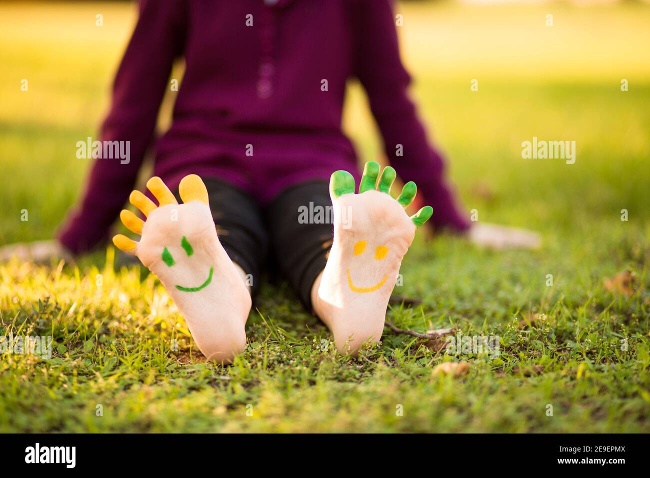 Happy feet con divertente vernice faccia all'aperto nel parco primaverile. Bambino sdraiato su erba verde. Concetto di ecologia Foto Stock