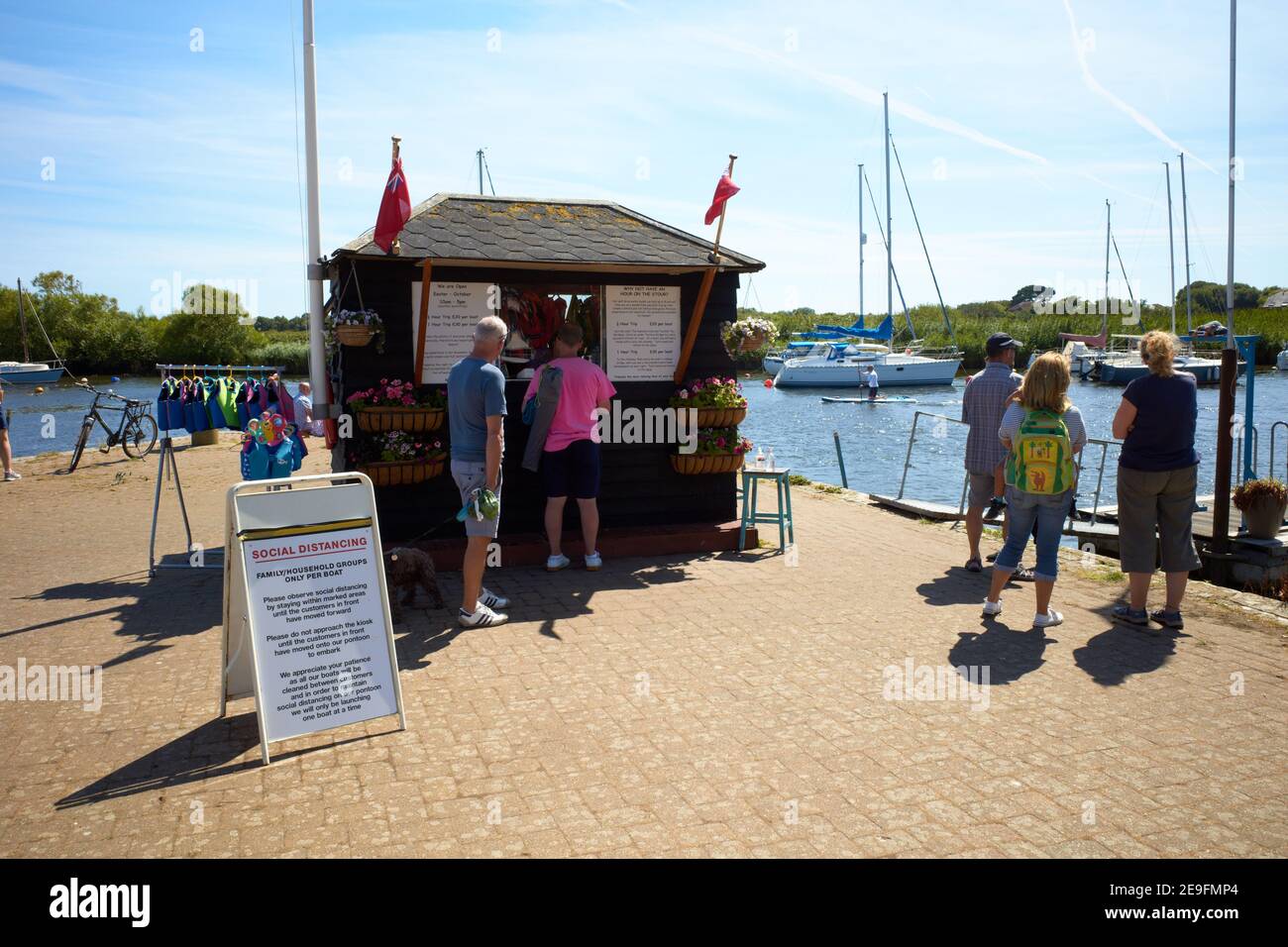 Fotografia di persone che si accodano per i biglietti per il noleggio di barche al porto di Christchurch, Devon, Regno Unito. Il cartello di allontanamento sociale è posizionato davanti alla biglietteria. Foto Stock