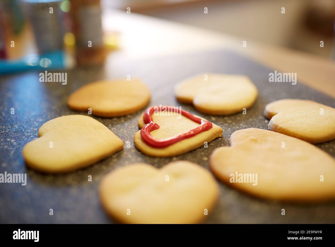Una fotografia ravvicinata di biscotti a forma di cuore su una superficie di lavoro della cucina. Un biscotto è in parte decorato con gli altri in procinto di essere decorato. Foto Stock