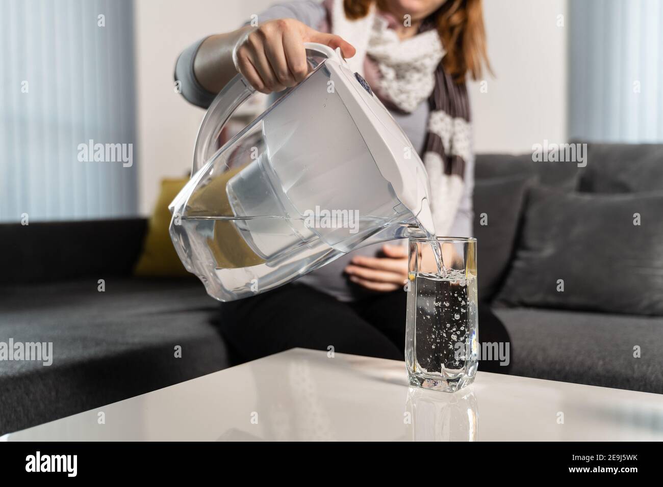 Donna sconosciuta che versa acqua filtrata in vetro sul tavolo dalla caraffa con filtro in plastica a casa durante il giorno - idratazione dell'acqua pulita concetto di vita sana Foto Stock
