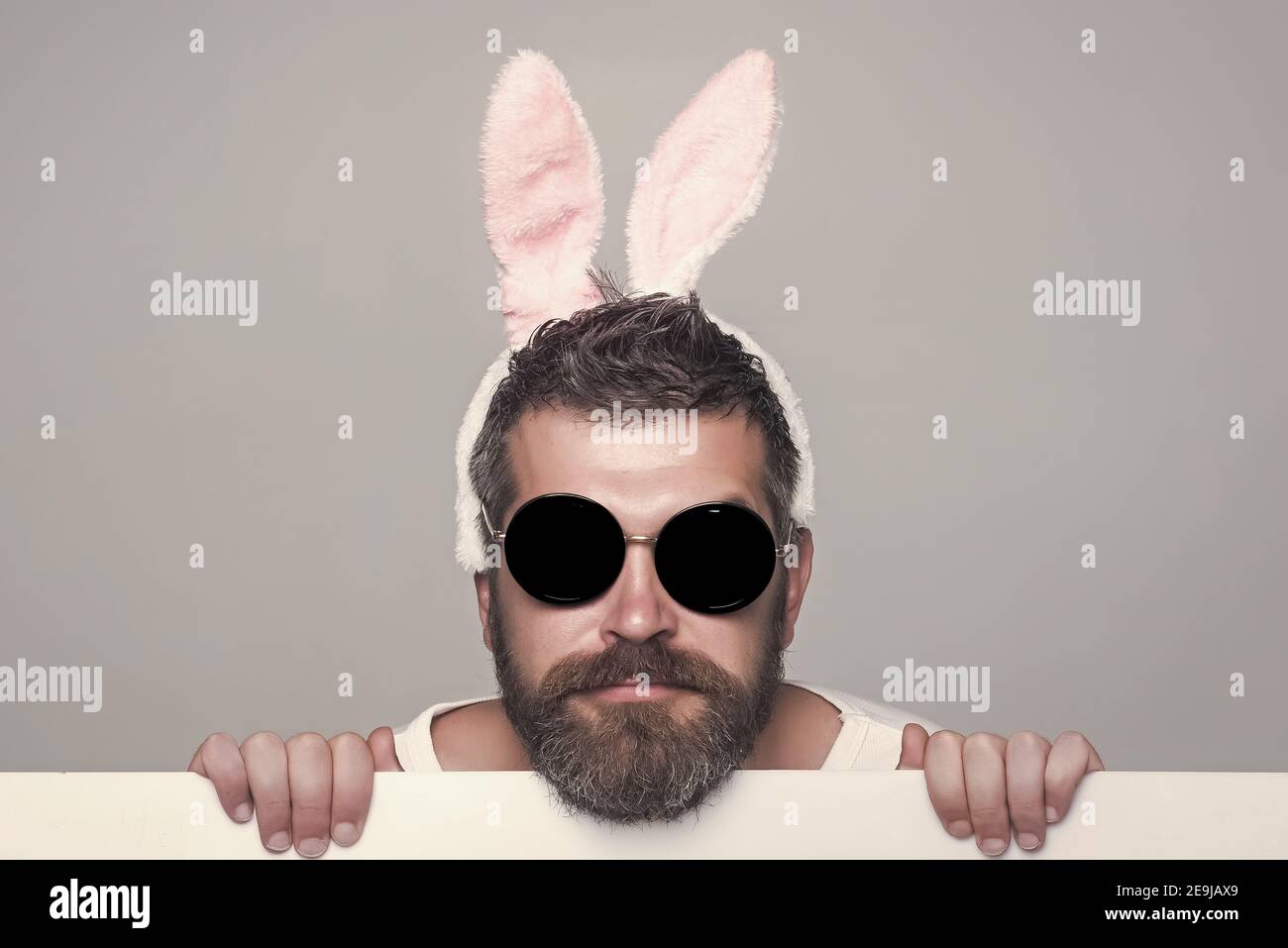 Uomo divertente con barba in orecchie conigliate e bicchieri su faccia grave con carta su sfondo grigio Foto Stock