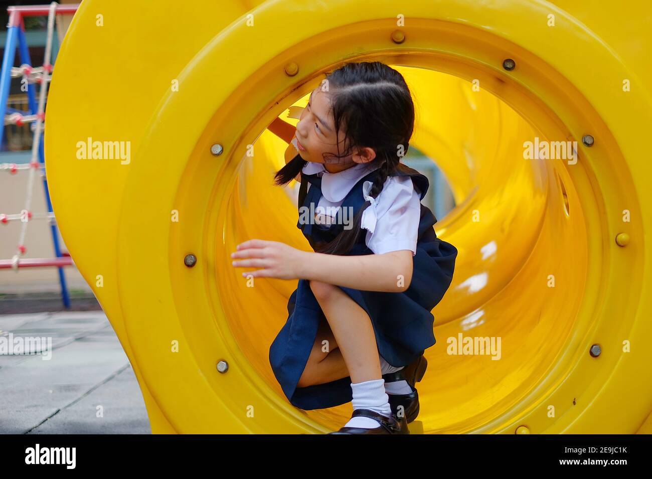Una giovane ragazza asiatica carina in bianco e blu scuola l'uniforme sta strisciando attraverso un tunnel di plastica giallo mentre gioca nascondi e cerca in un parco giochi dopo Foto Stock