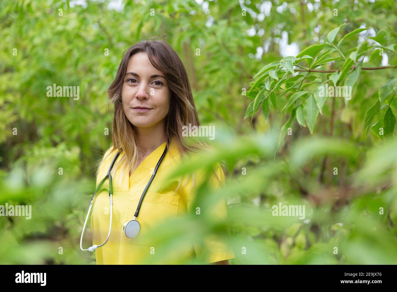 Ritratto di giovane infermiera vestita in uniforme con stetoscopio. Sorride in un ambiente naturale circondato da alberi, piante e vege Foto Stock