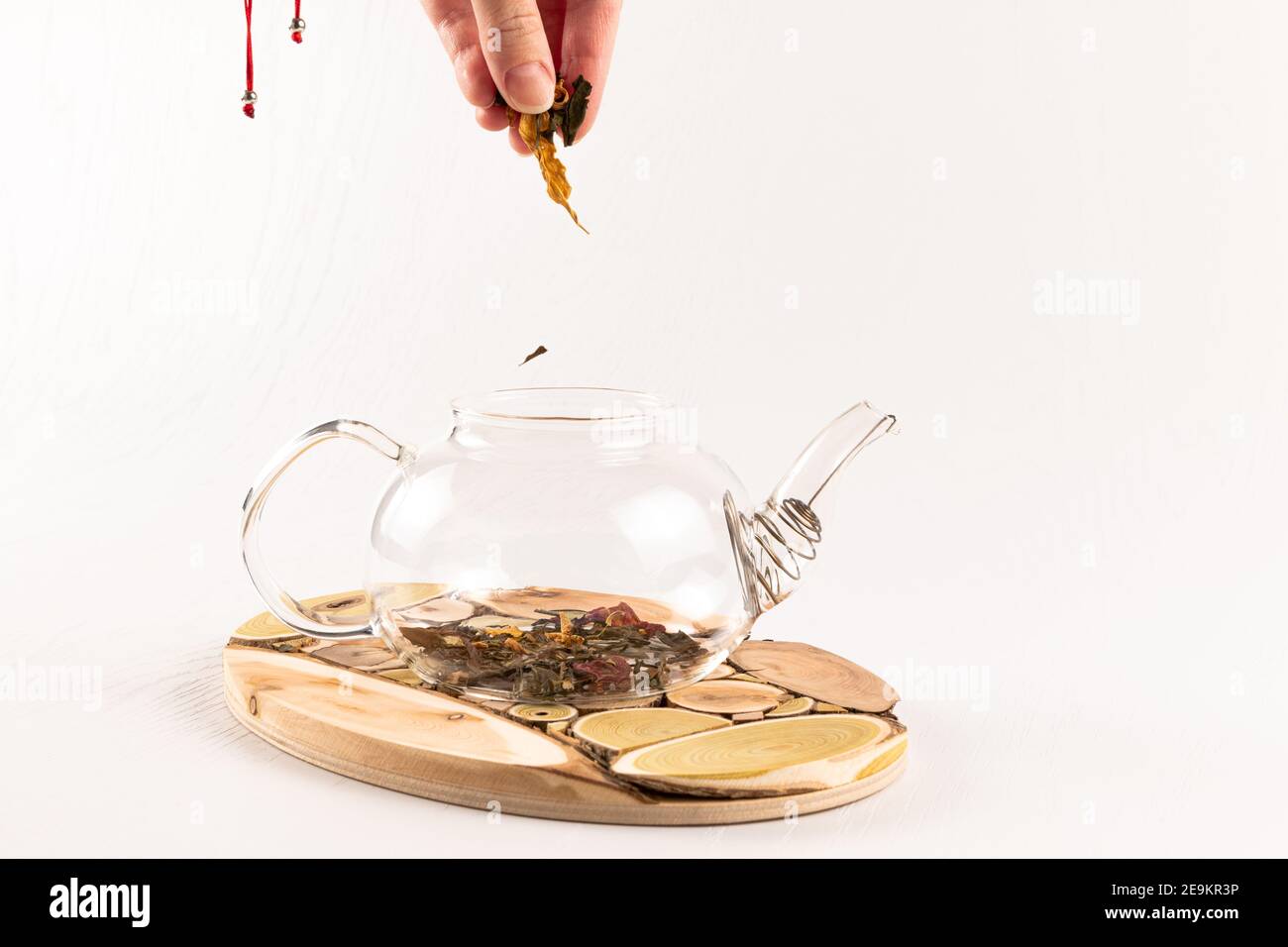 una mano femminile versa il tè alle erbe aromatiche cinesi in un teiera in vetro su un supporto in legno su un bianco tavolo di legno Foto Stock