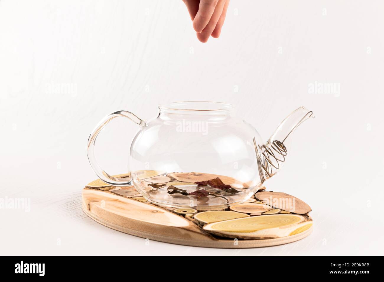 una mano femminile versa il tè alle erbe aromatiche cinesi in un teiera in vetro su un supporto in legno su un bianco tavolo di legno Foto Stock