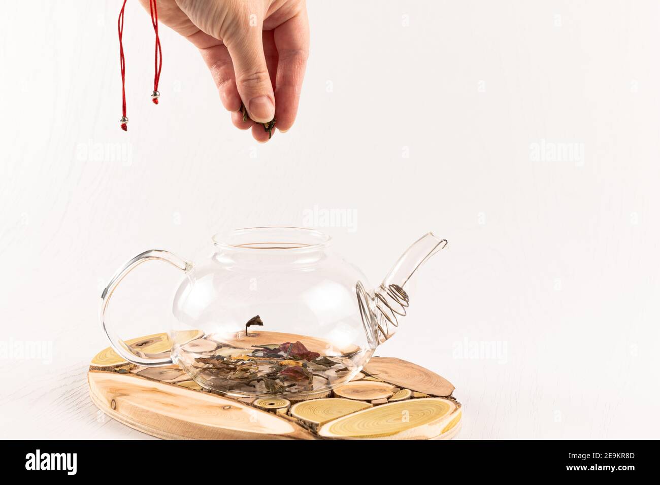 una mano femminile versa il tè alle erbe aromatiche cinesi in un teiera in vetro su un supporto in legno su un bianco tavolo di legno Foto Stock