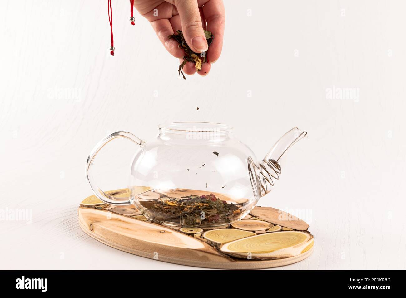 una mano femminile versa il tè alle erbe aromatiche cinesi in un teiera in vetro su un supporto in legno su un bianco tavolo di legno Foto Stock