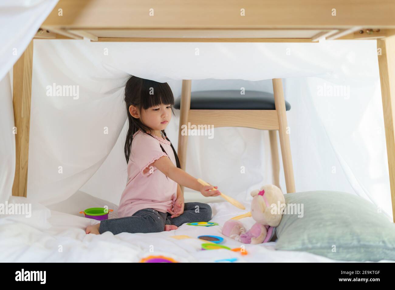 Asian cute bambina giocare cucina e alimentare cibo a. la sua bambola mentre si siede in una fortezza coperta in vita camera a casa per un perfetto nascondiglio lontano da Foto Stock