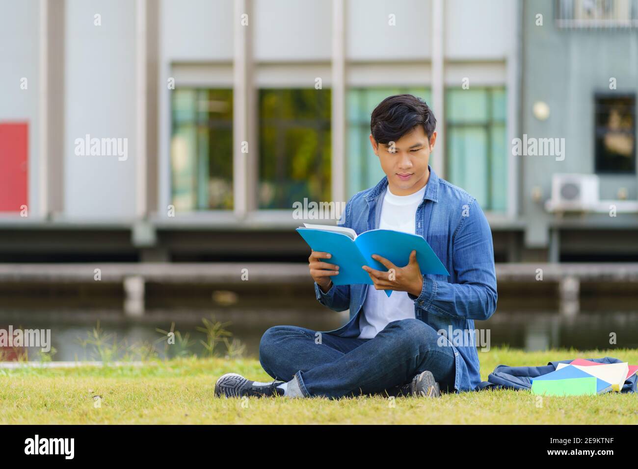 Ritratto dell'uomo asiatico studente universitario aitting su erba nel campus cercando felice e la lettura di un libro nel parco. Foto Stock