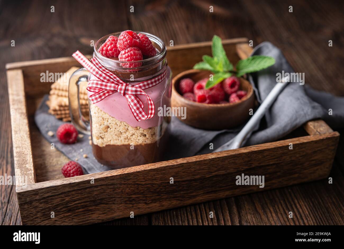 Dessert a strati senza cottura con mousse al cioccolato fondente, biscotti schiacciati e crema di lamponi in vaso di vetro su rustico fondo di legno Foto Stock